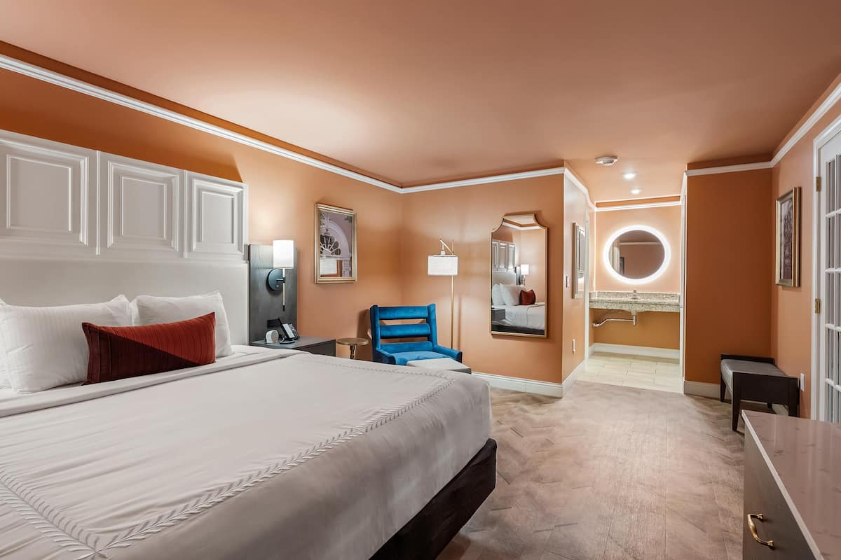 A hotel room with a large bed, a blue chair, a vanity with a mirror, and a round light fixture.
