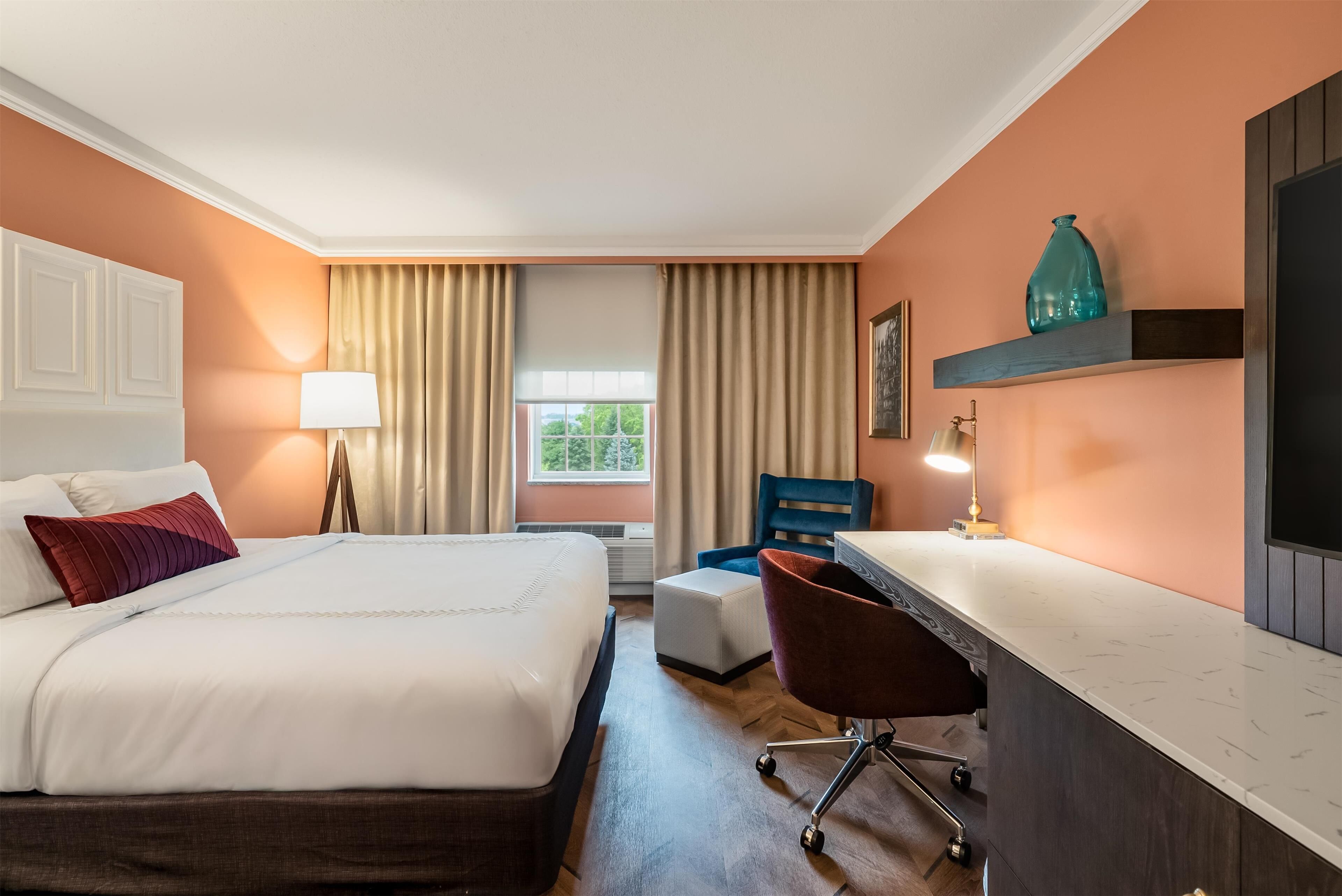 A hotel room with a bed, a desk, a chair, a lamp, a window with curtains, and a shelf with a vase.