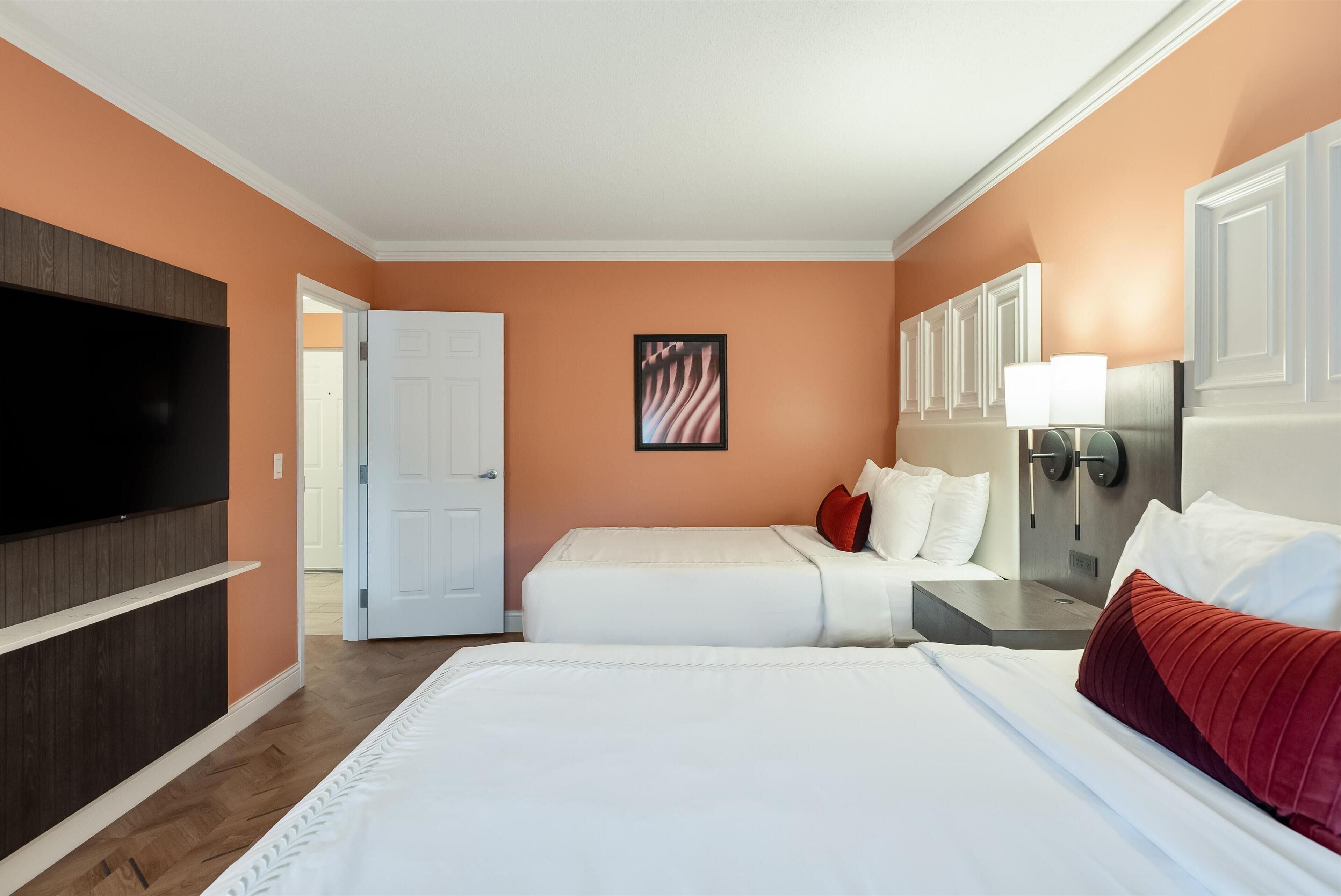 A hotel room with a large flat-screen TV, a bed with white bedding, a bedside table with a lamp, and a door leading to another room.