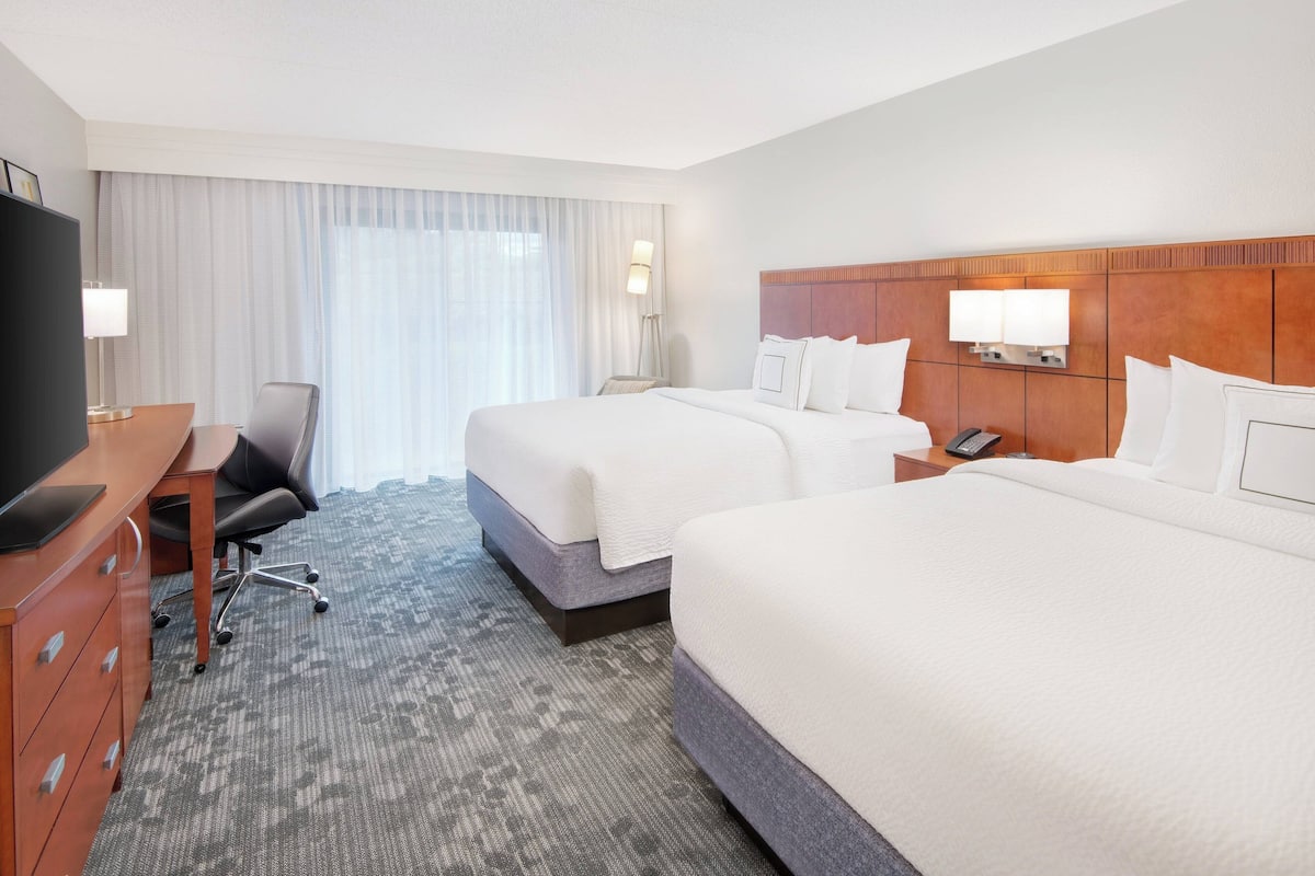 Een hotelkamer met twee bedden, een bureau met stoel, een televisie en een groot raam met gordijnen.
