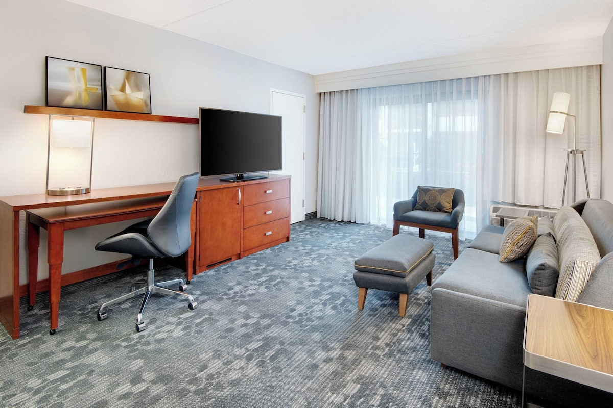 Een hotelkamer met een houten bureau, een flatscreen televisie, een grijze bank, een poef en een groot raam met dunne gordijnen.