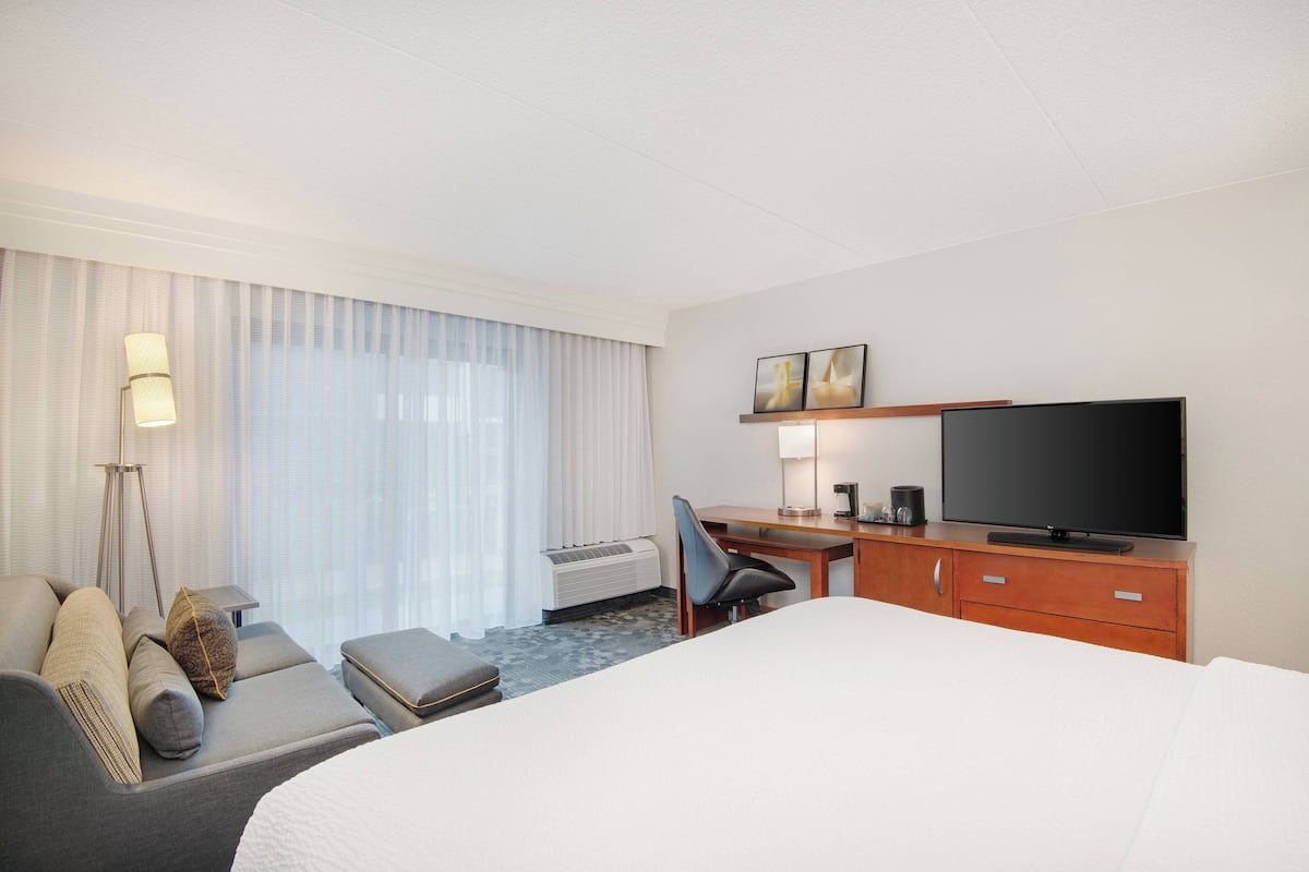Een hotelkamer met een bed, een bureau met een televisie, een stoel, een lamp en een groot raam met gordijnen.
