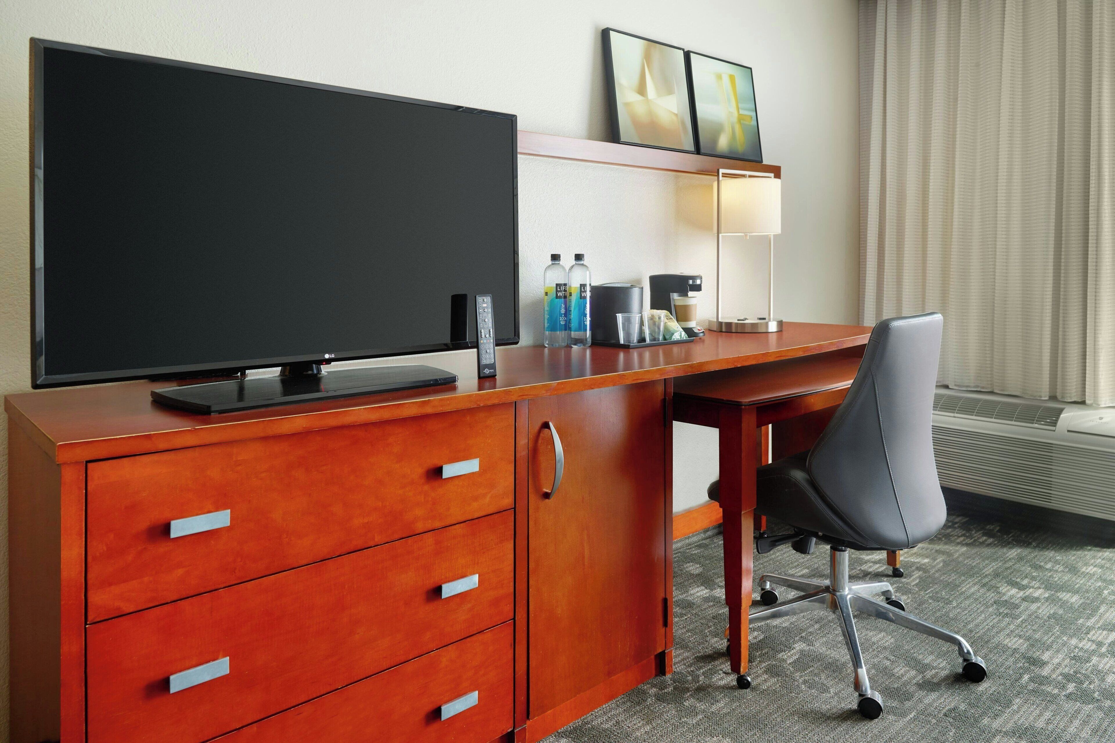 A hotel room with a wooden desk, a flat-screen TV, a chair, a lamp, and a window with curtains.