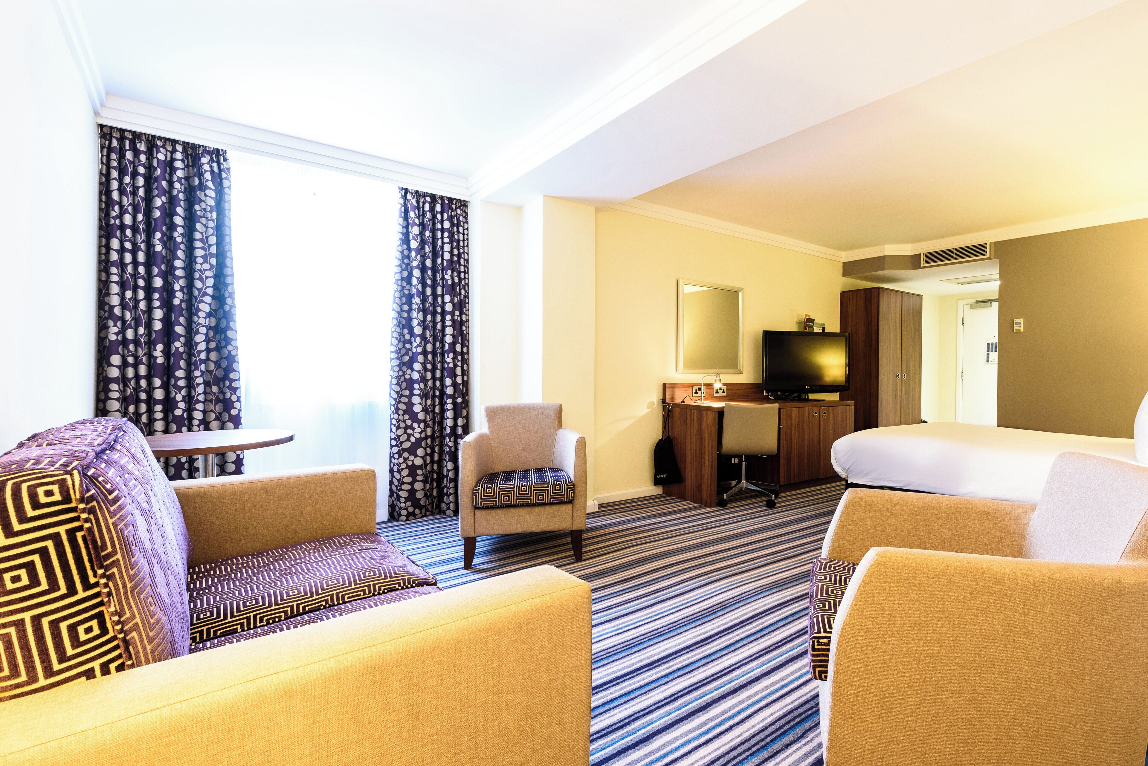 A hotel room with a sofa, armchair, and a bed. There is a desk with a chair and a television.