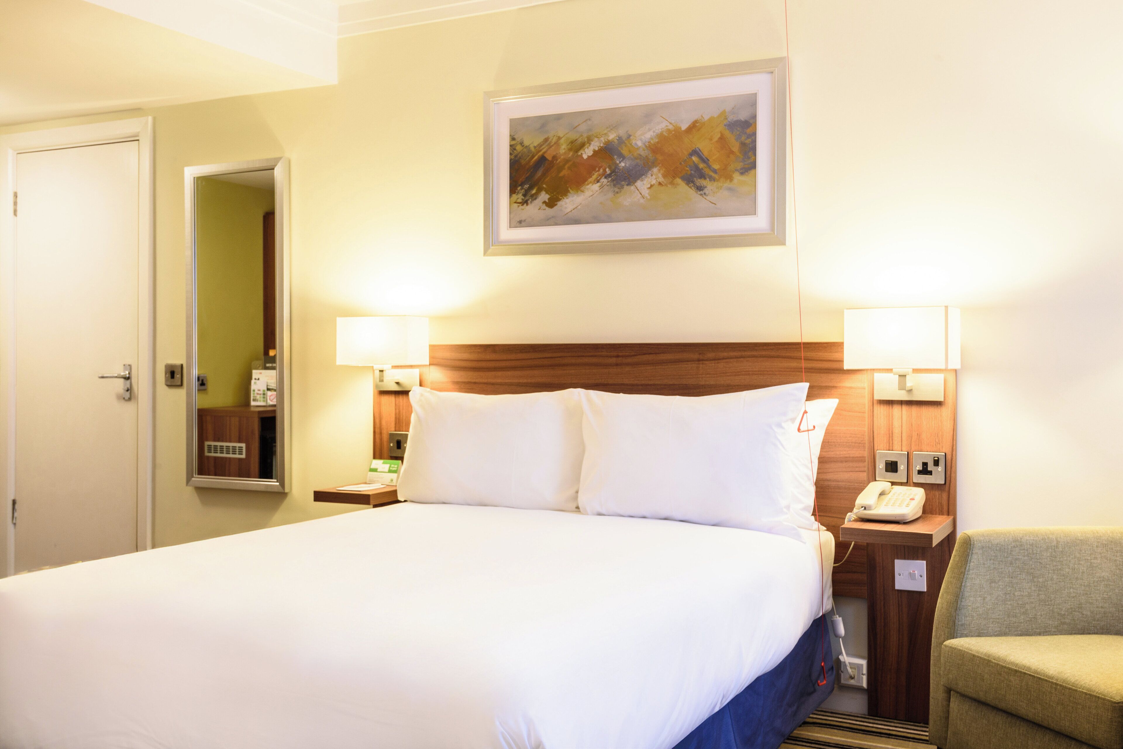 A hotel room with a large bed, two bedside lamps, a wooden headboard, a painting on the wall, a bedside telephone, and a chair.