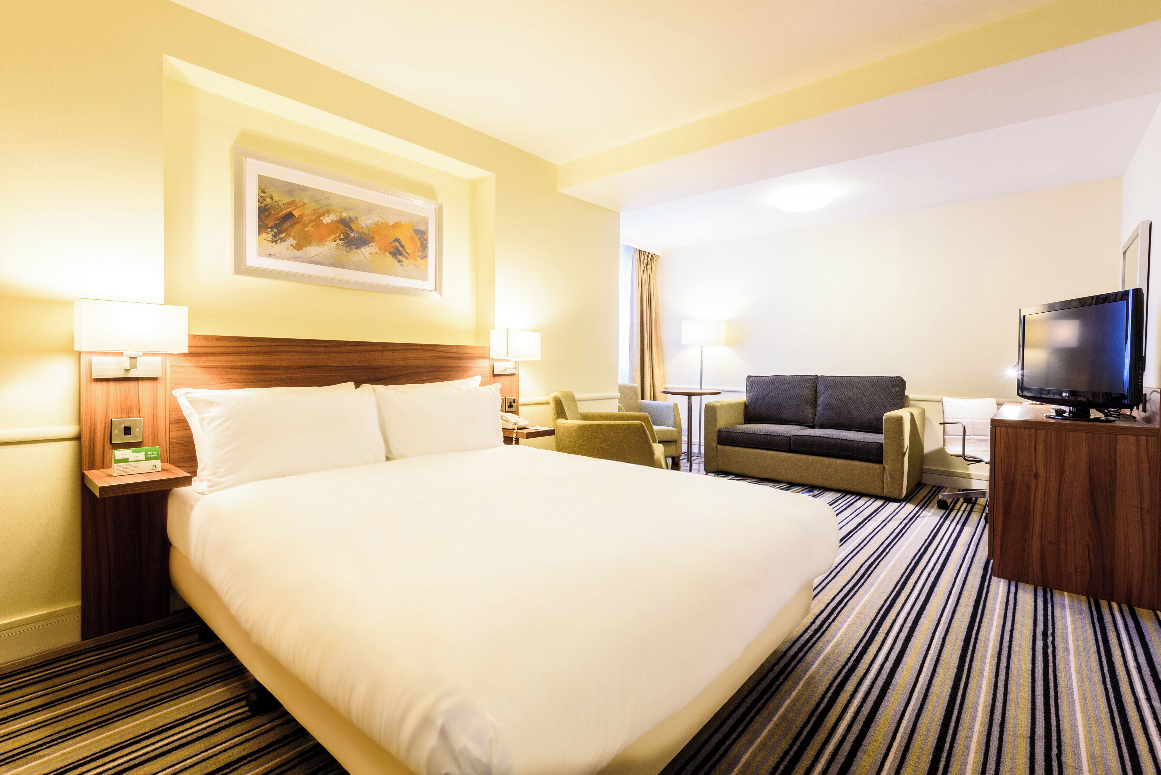 A hotel room with a large bed, a sofa, a television, and a painting on the wall.
