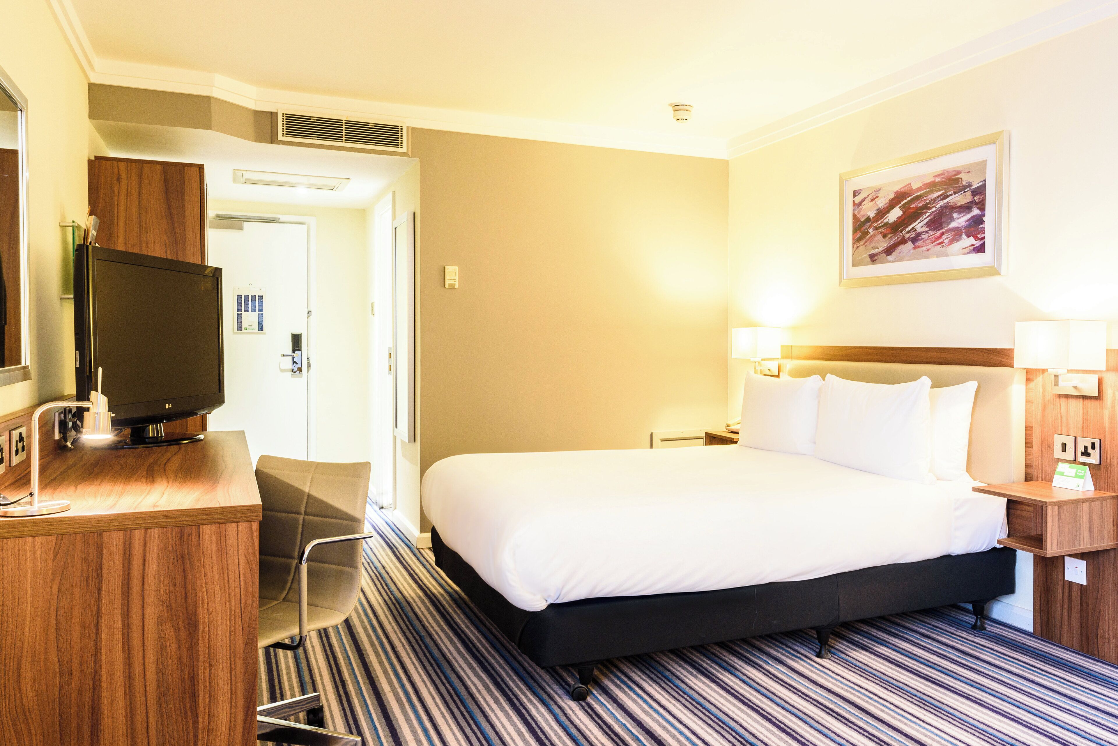 A hotel room with a bed, a desk with a television, a chair, and a painting on the wall.