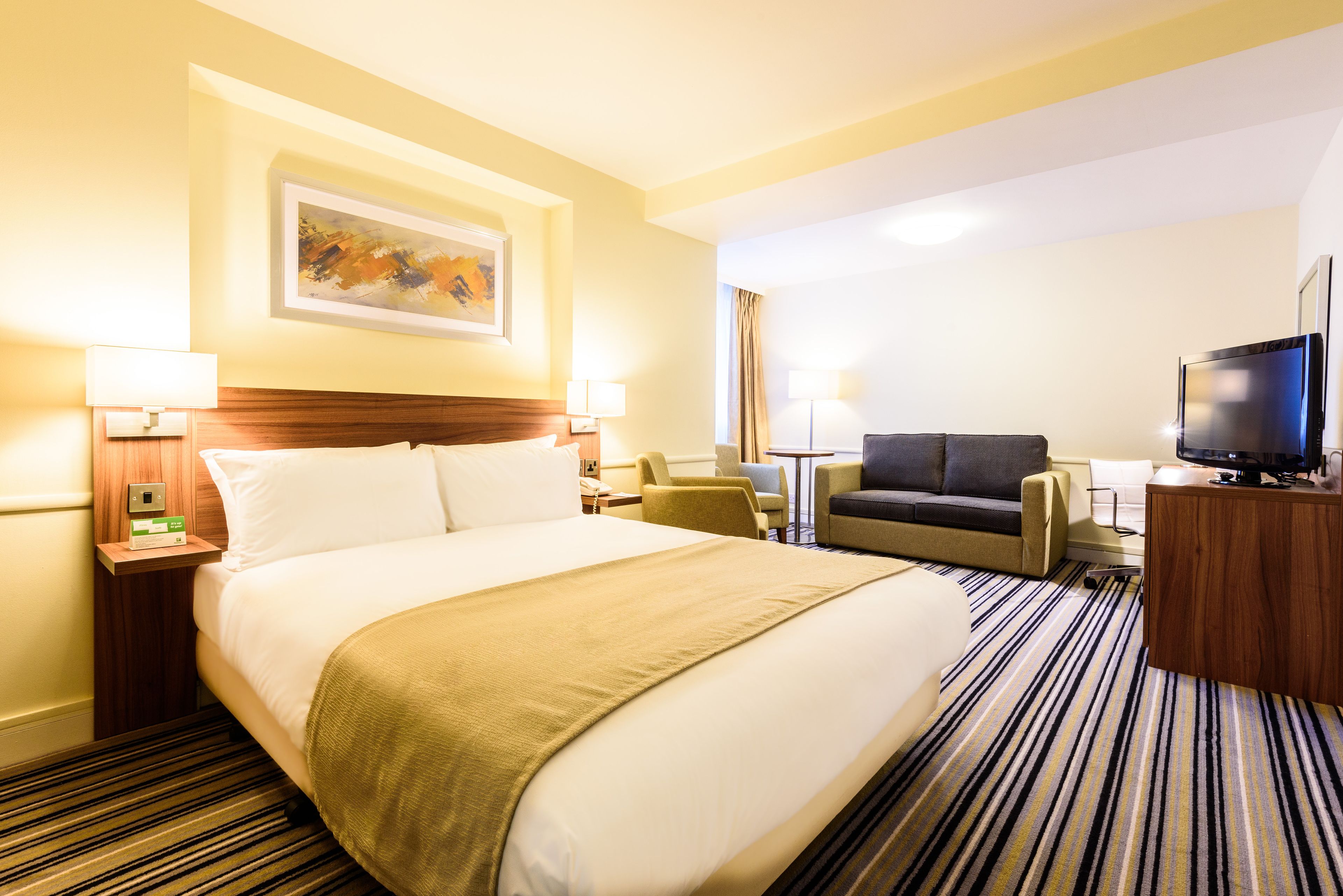A hotel room with a large bed, a desk with a TV, a sofa, and a painting on the wall.