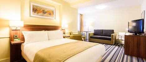 A hotel room with a large bed, a desk with a TV, a sofa, and a painting on the wall.