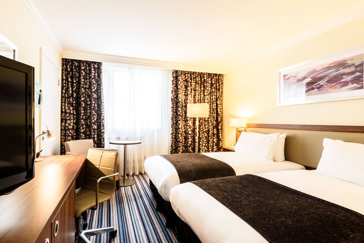 Ein Hotelzimmer mit zwei Betten, einem Schreibtisch, einem Sessel, einem Fernseher und einem Fenster mit Vorhängen.
