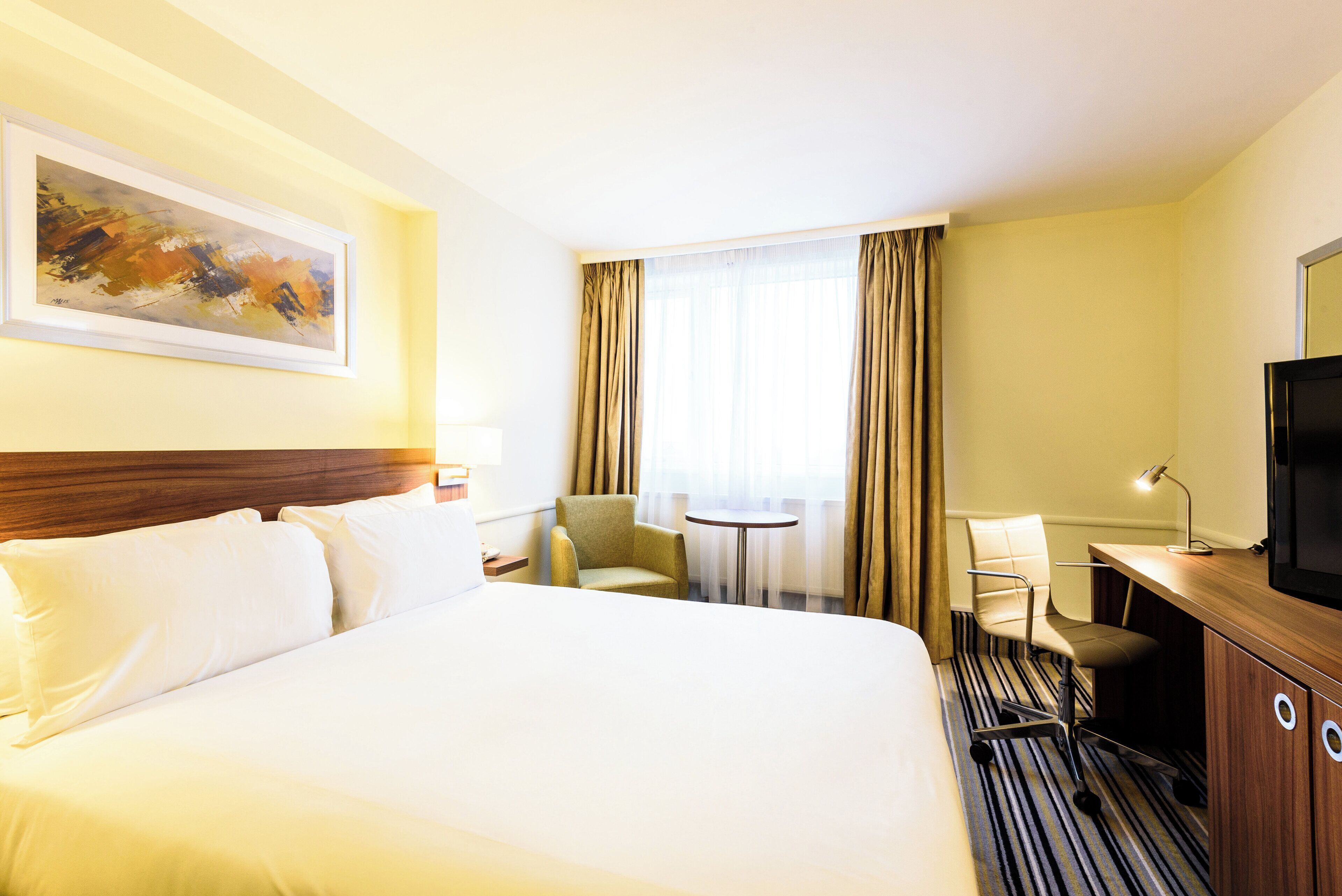 A hotel room with a large bed, a desk with a TV, a chair, a small table, and a painting on the wall.