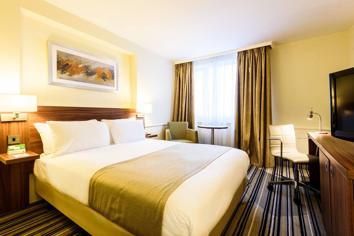 Ein Hotelzimmer mit einem großen Bett, einem Schreibtisch mit Fernseher, einem Sessel, einem kleinen Tisch und einem Gemälde an der Wand.