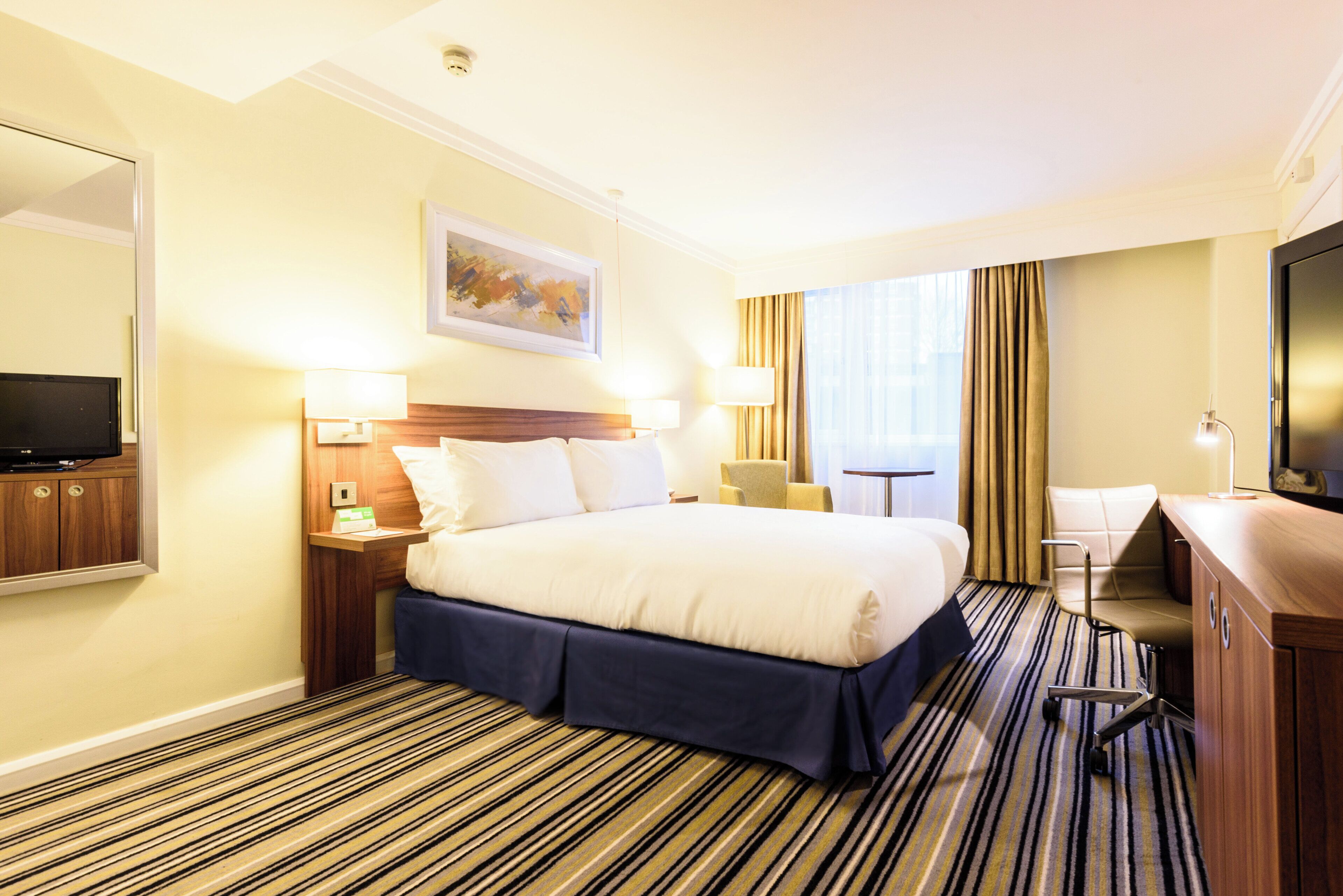 A hotel room with a large bed, a television, a desk with a lamp, and a window with curtains.