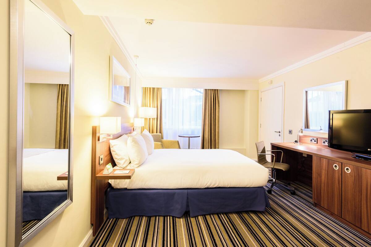 Ein Hotelzimmer mit Bett, Nachttisch, Lampe, Fernseher und einer kleinen Schreibtischzeile.
