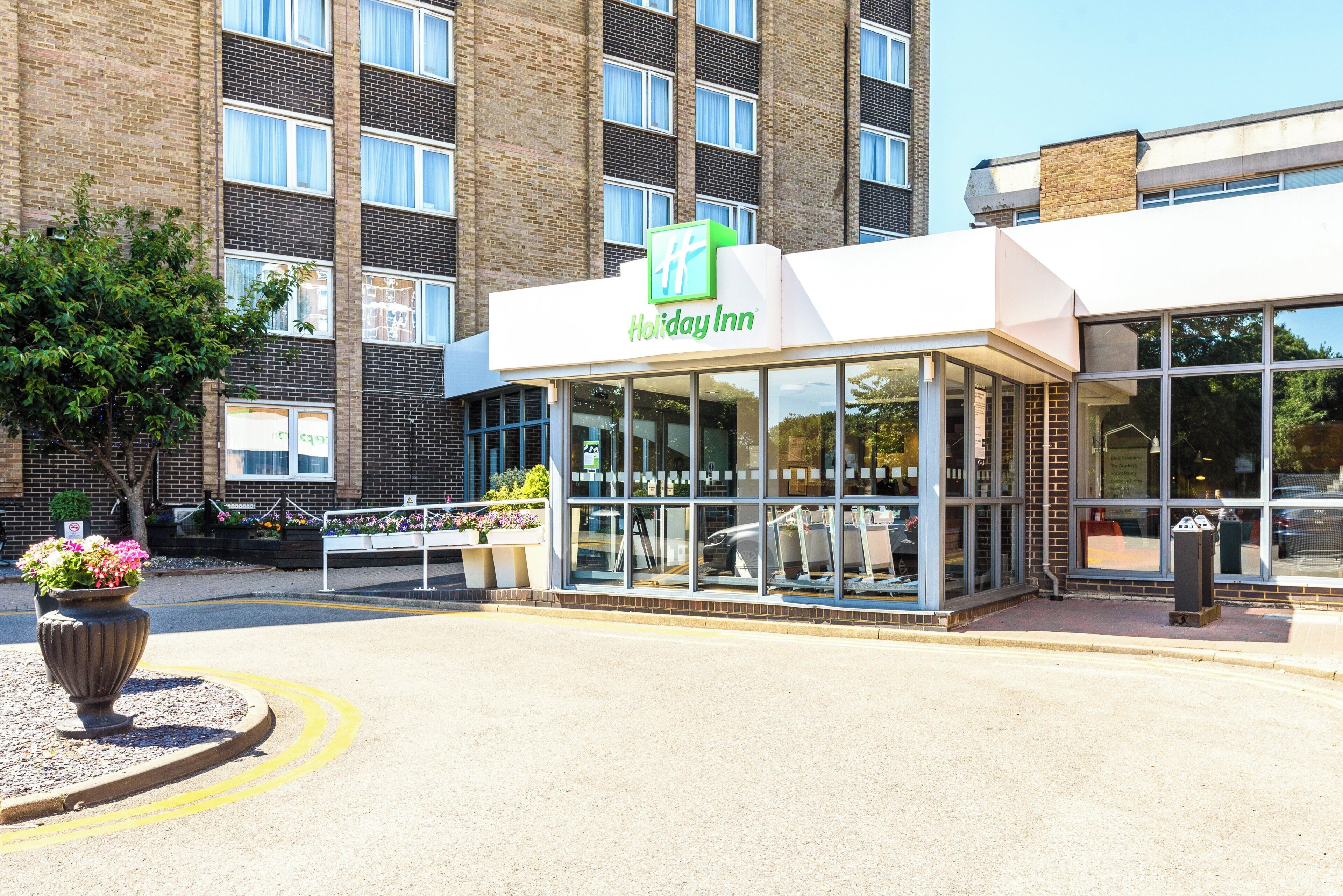 Un hotel Holiday Inn con fachada de vidrio moderna, área de asientos al aire libre y un edificio de ladrillo al fondo.