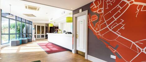 Un vestíbulo de hotel con una recepción, un ventanal grande y una pared roja decorada con un diseño de mapa.