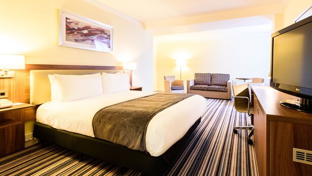 Quarto de hotel com cama, escrivaninha, televisão e uma cadeira.