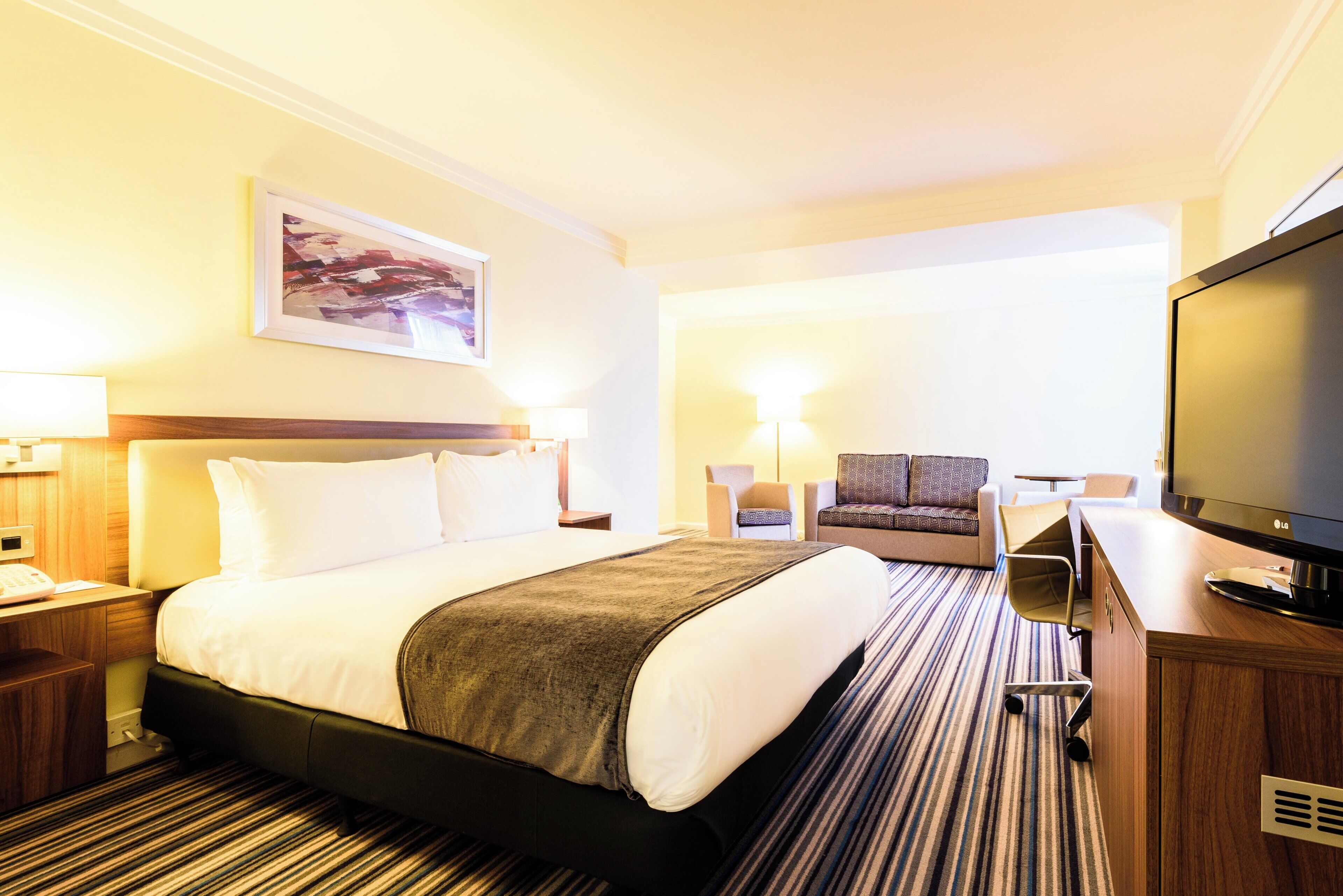 Una habitación de hotel con una cama, un escritorio, una silla, un televisor y un cuadro en la pared.