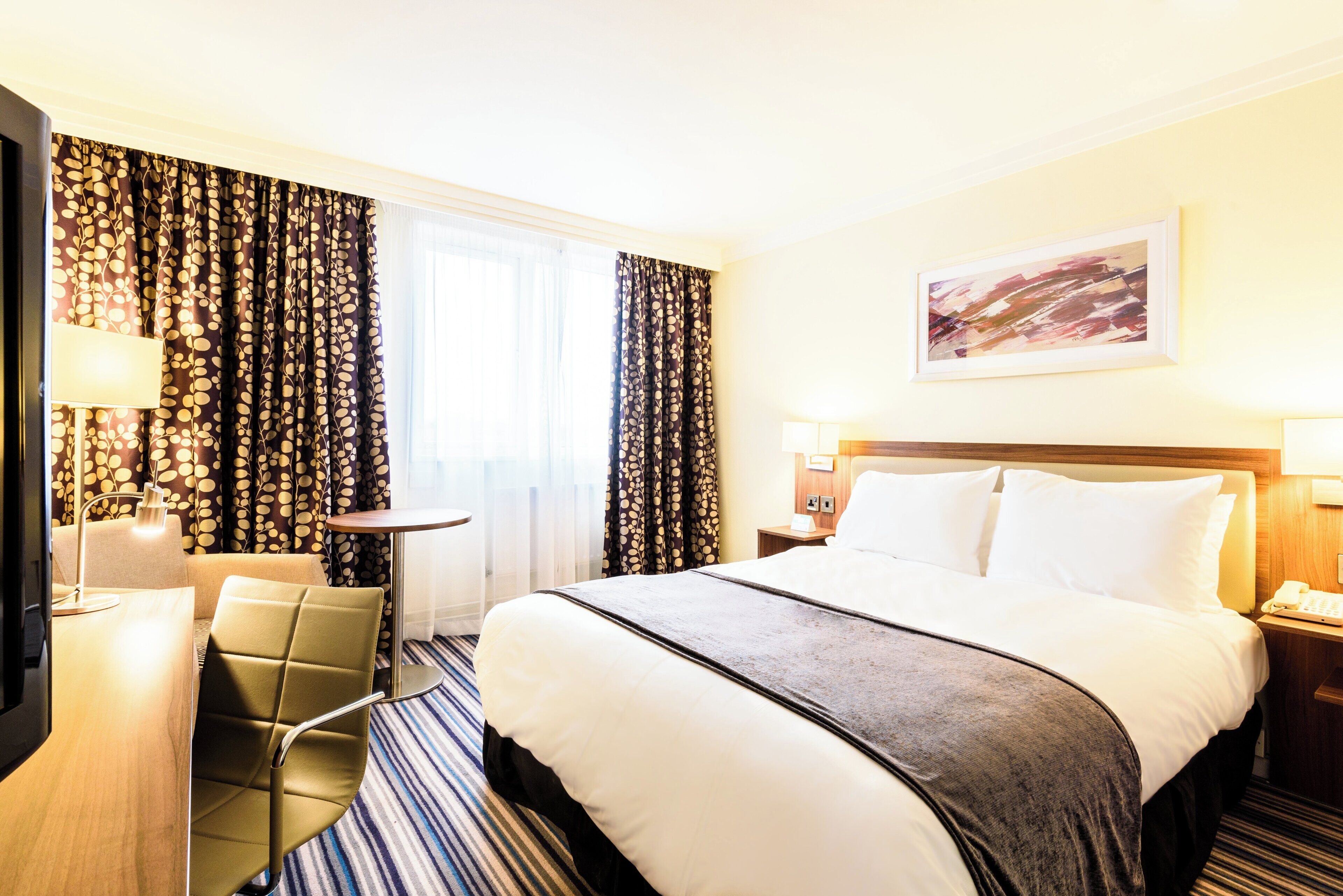 A hotel room with a bed, a desk, a chair, a lamp, and a painting on the wall.