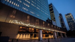 Um hotel moderno com fachada de vidro proeminente, entrada bem iluminada e vista clara dos edifícios altos ao redor.