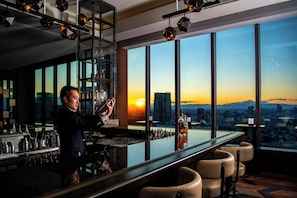 A bar with a city view at sunset, a bartender, and a high-rise building in the background.