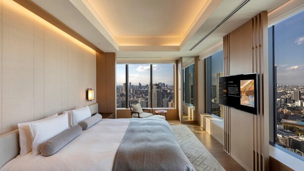 Quarto de hotel moderno com uma cama grande, televisão, vista da cidade e uma pequena mesa com uma cadeira.