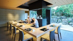 Cozinha moderna com chefs preparando comida, uma longa mesa de jantar com cadeiras e vista para um jardim rochoso.