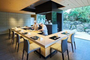 A modern kitchen with chefs preparing food, a long dining table with chairs, and a view of a rocky garden. - Hotel Okura Tokyo South Wing (Tokyo)