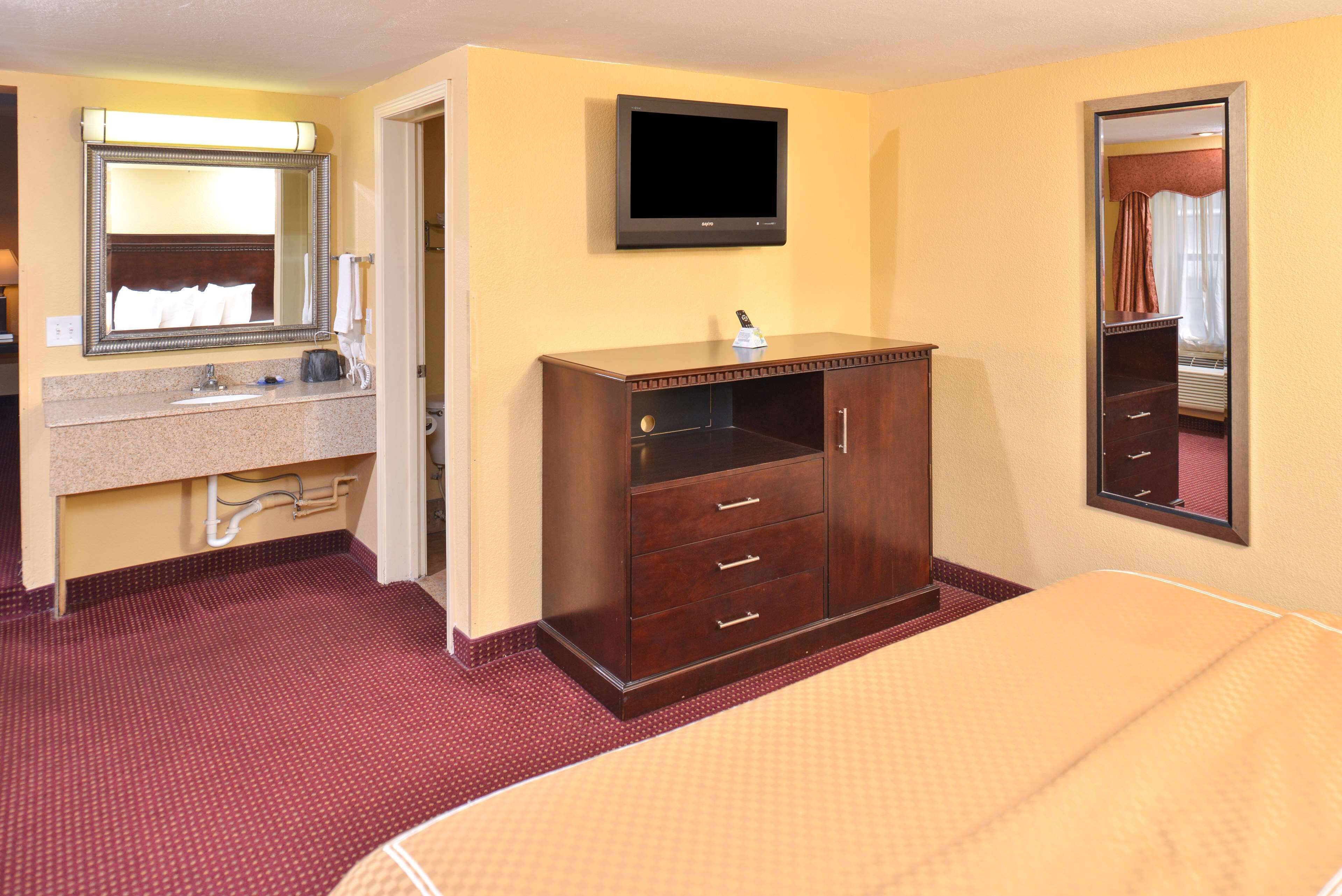 A hotel room with a bed, a wooden dresser, a television mounted on the wall, and a bathroom with a sink and mirror.