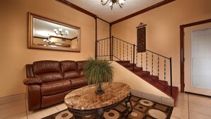 A lobby with a leather sofa, a round wooden coffee table, a staircase with a red carpet, and a large mirror on the wall.