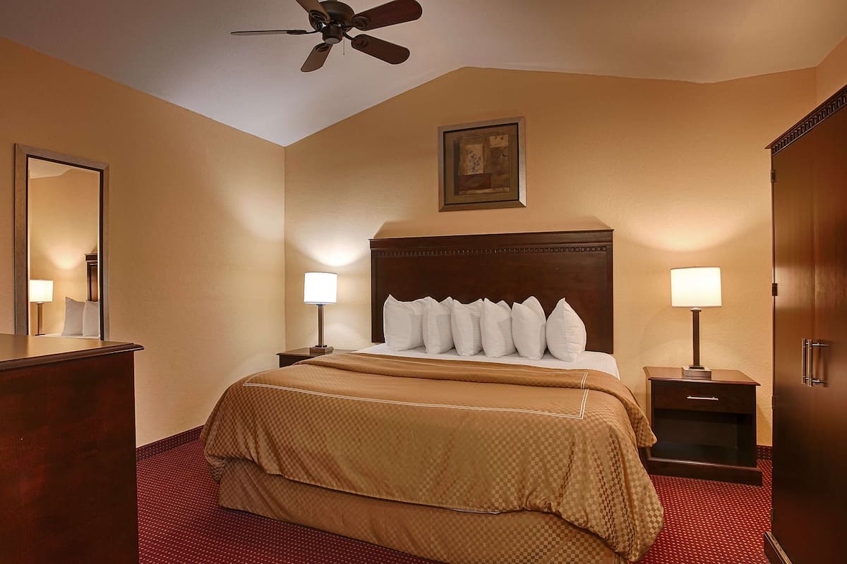 A hotel room with a large bed, two bedside tables with lamps, a wooden headboard, a ceiling fan, a framed picture on the wall, and a red carpeted floor.