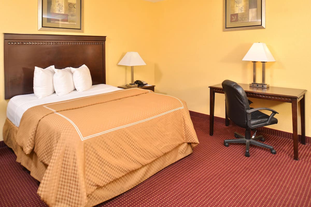 A hotel room with a bed, two bedside tables with lamps, a desk with a chair, and a red carpet.