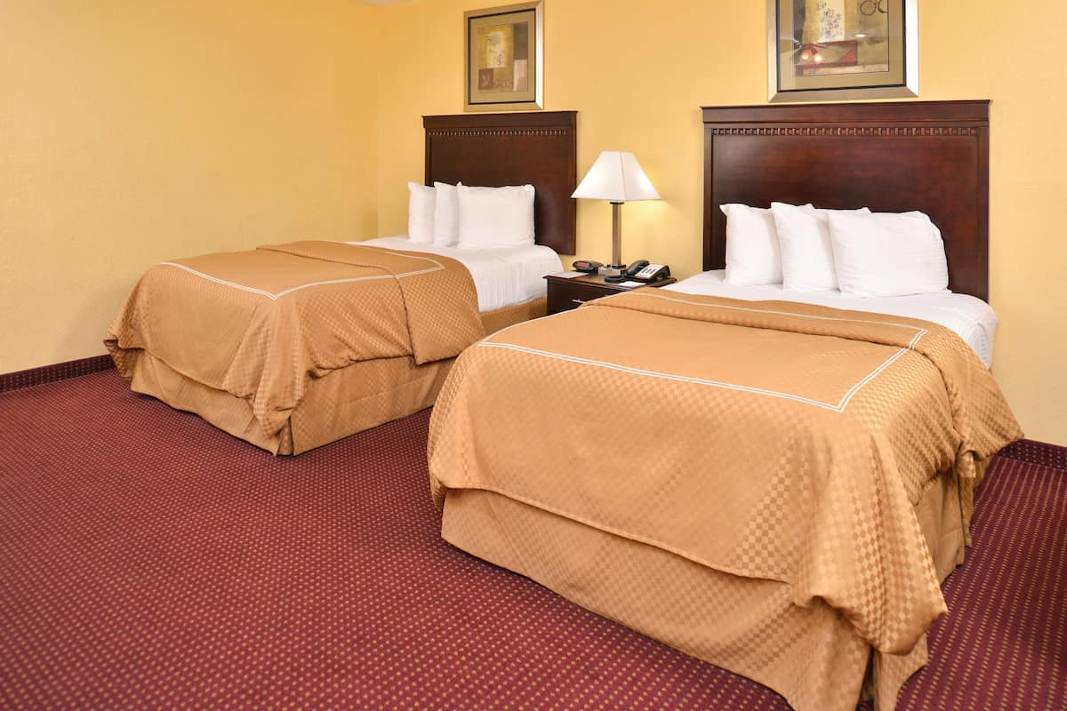 A hotel room with two beds, each with a brown bedspread and white pillows, a wooden headboard, a nightstand with a lamp, and a framed picture on the wall.