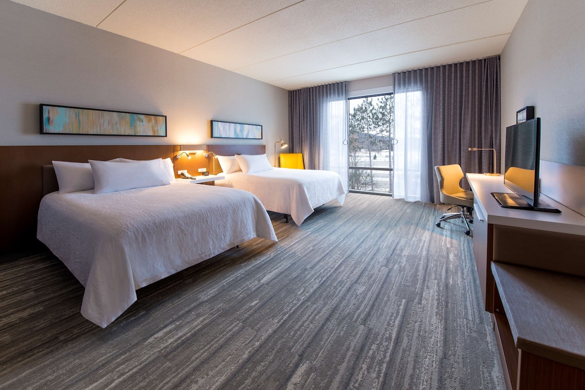 Une chambre d’hôtel avec deux lits, un bureau, une chaise et une grande fenêtre avec des rideaux.