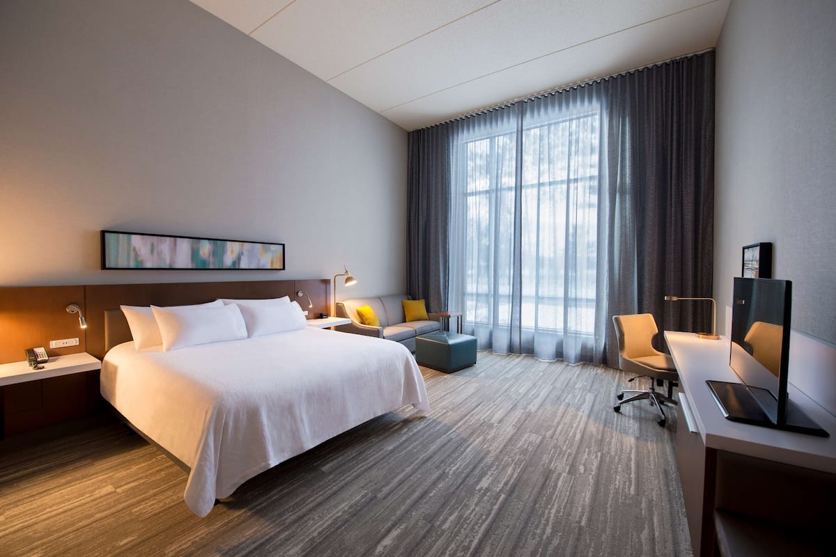 Une chambre d’hôtel avec un grand lit, un bureau avec un ordinateur, un canapé et une grande fenêtre avec des rideaux.