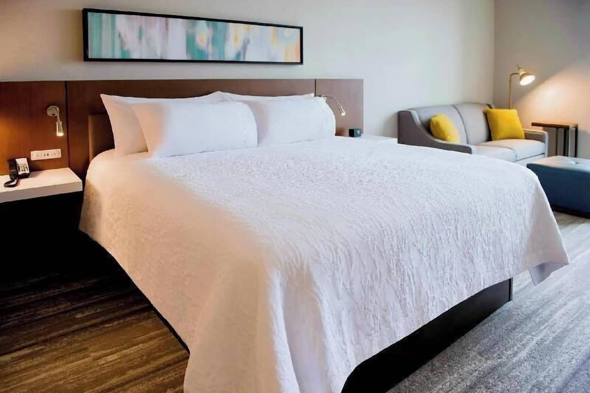 Une chambre d’hôtel avec un grand lit, une table de chevet avec une lampe, une petite table avec une lampe et un canapé avec des coussins.