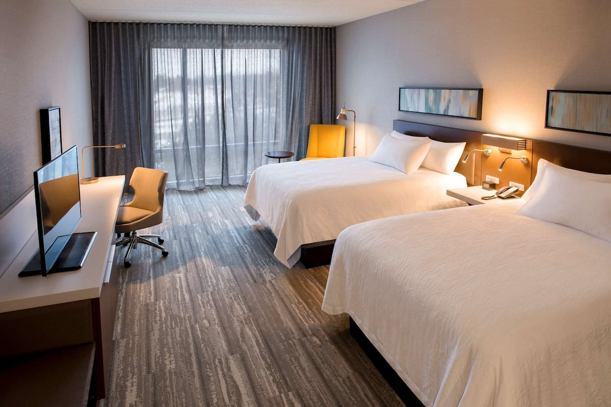 Une chambre d’hôtel avec deux lits, un bureau avec un ordinateur, une chaise, une lampe et une fenêtre avec des rideaux.