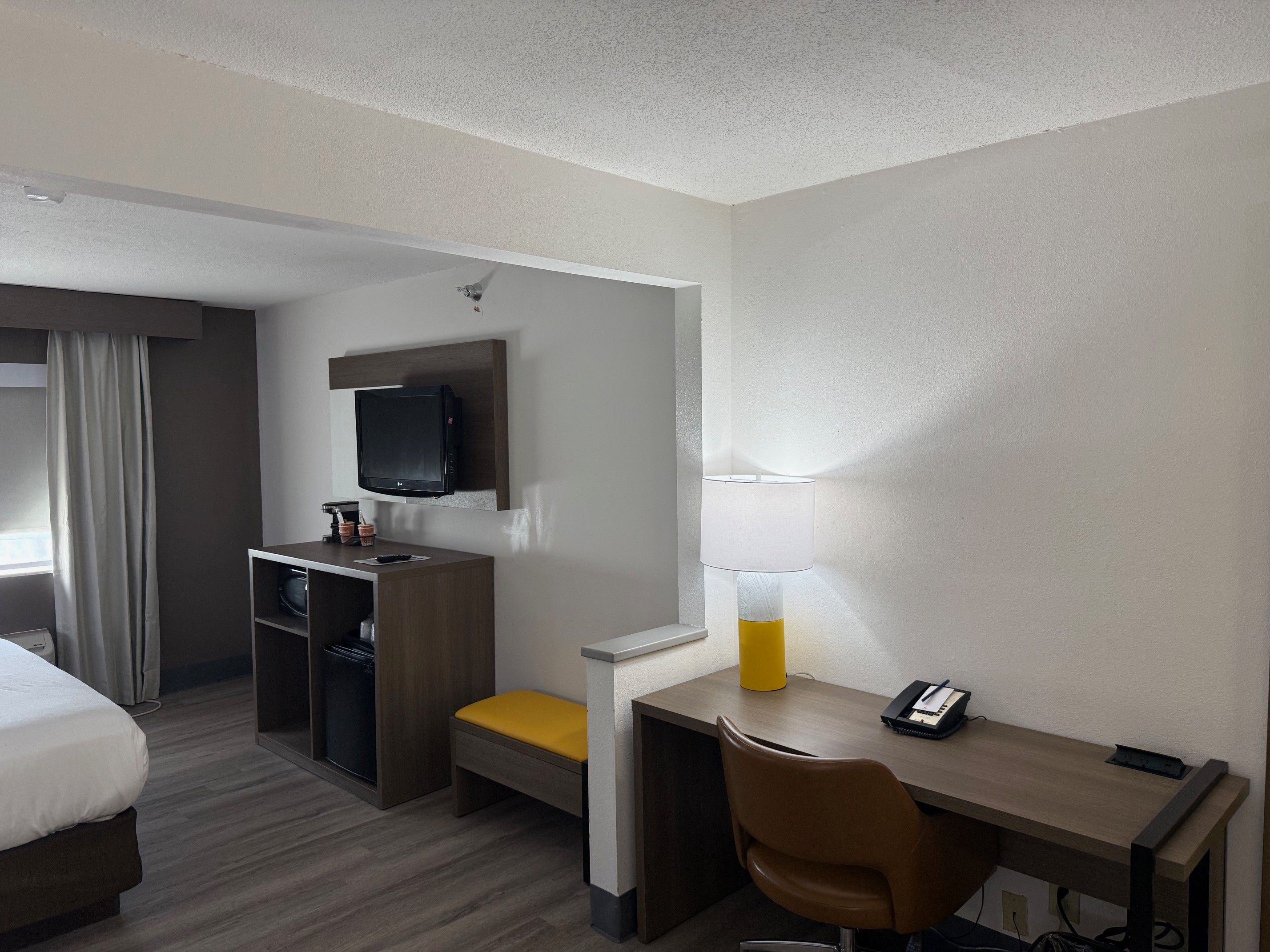 A hotel room with a bed, a desk, a chair, a television, a lamp, and a small table with a phone and a notebook.