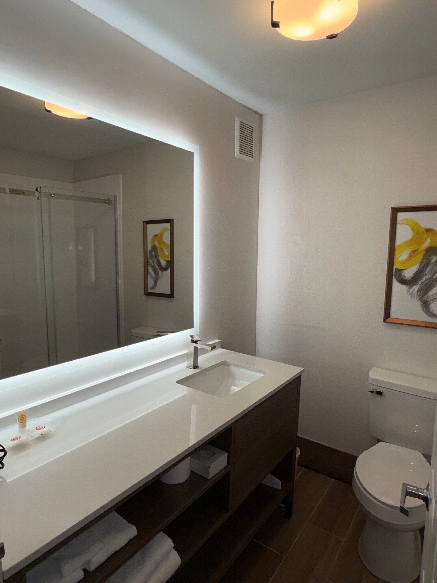 A bathroom with a white sink, mirror, and toilet. There is a towel rack and a framed artwork on the wall.