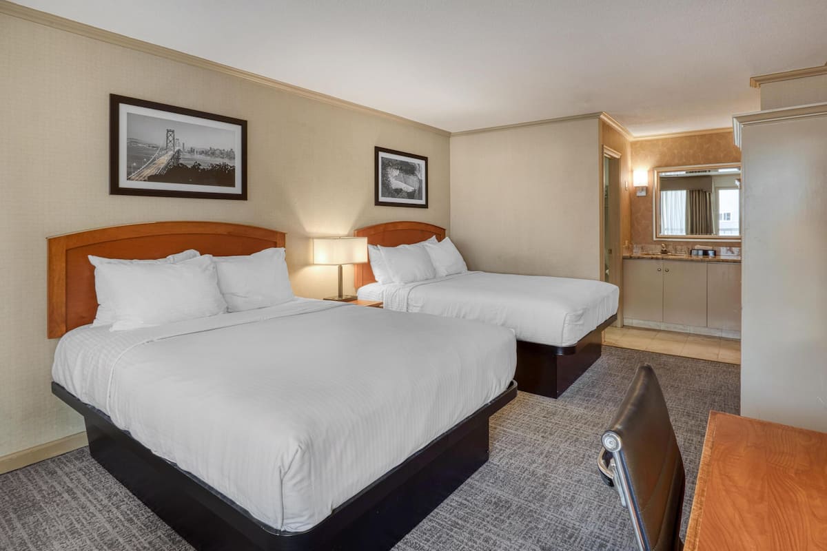 A hotel room with two beds, a desk, a chair, a mirror, and two framed pictures on the wall.
