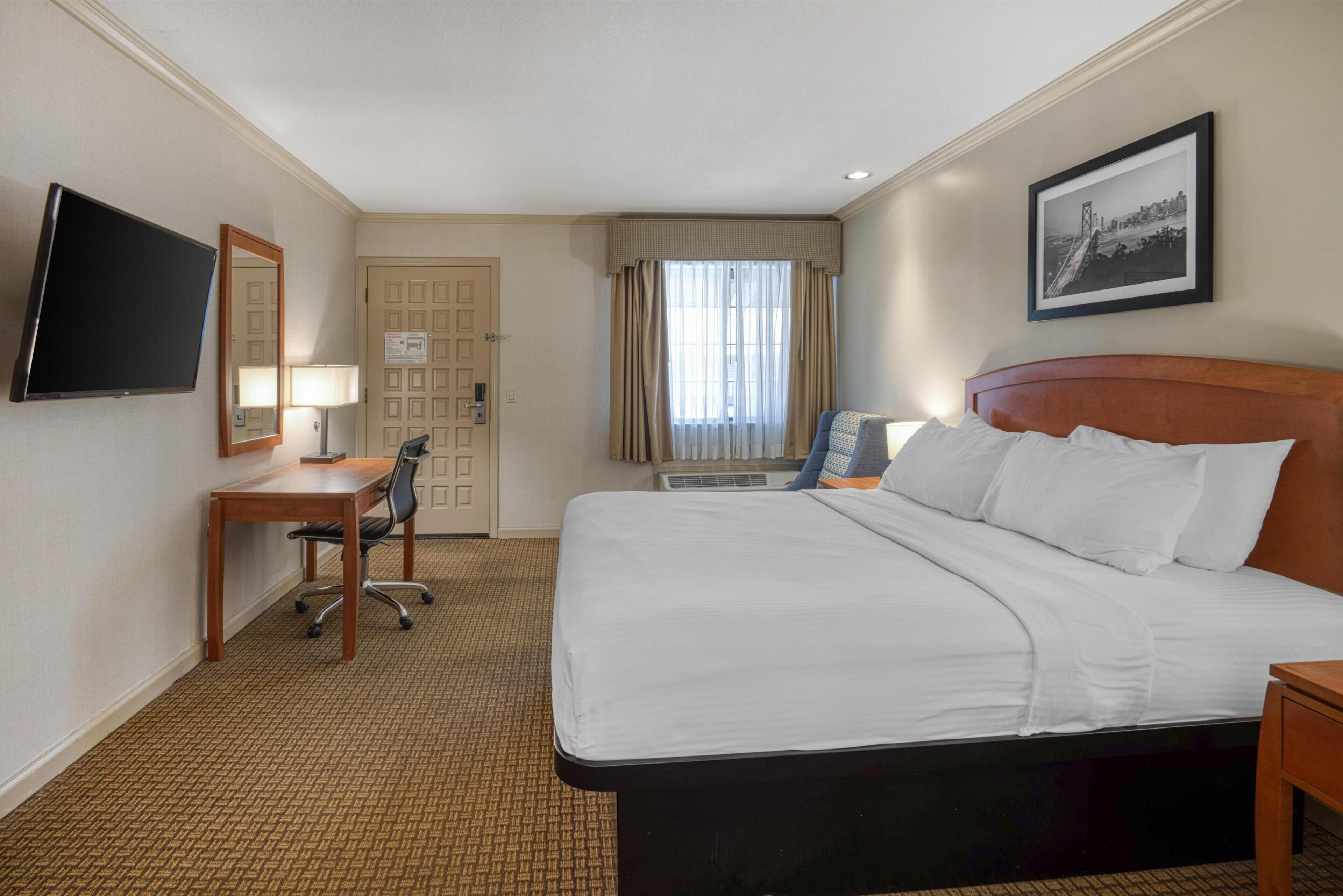 A hotel room with a bed, a desk with a chair, a television, a window with curtains, and a framed picture on the wall.
