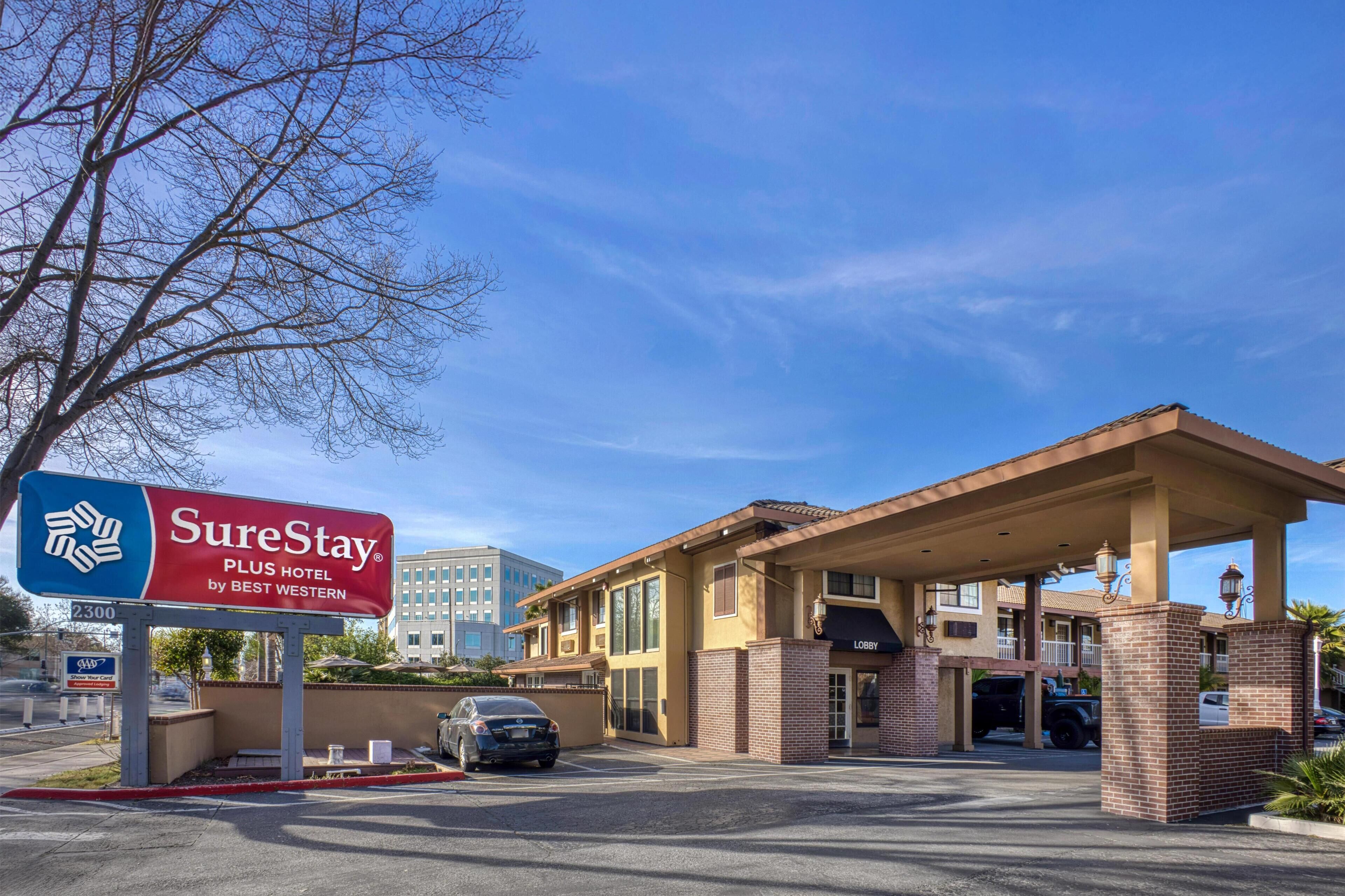 L’entrée d’un hôtel, avec une enseigne « SureStay Plus Hotel », une zone de stationnement couverte et un bâtiment à plusieurs étages doté de nombreuses fenêtres.