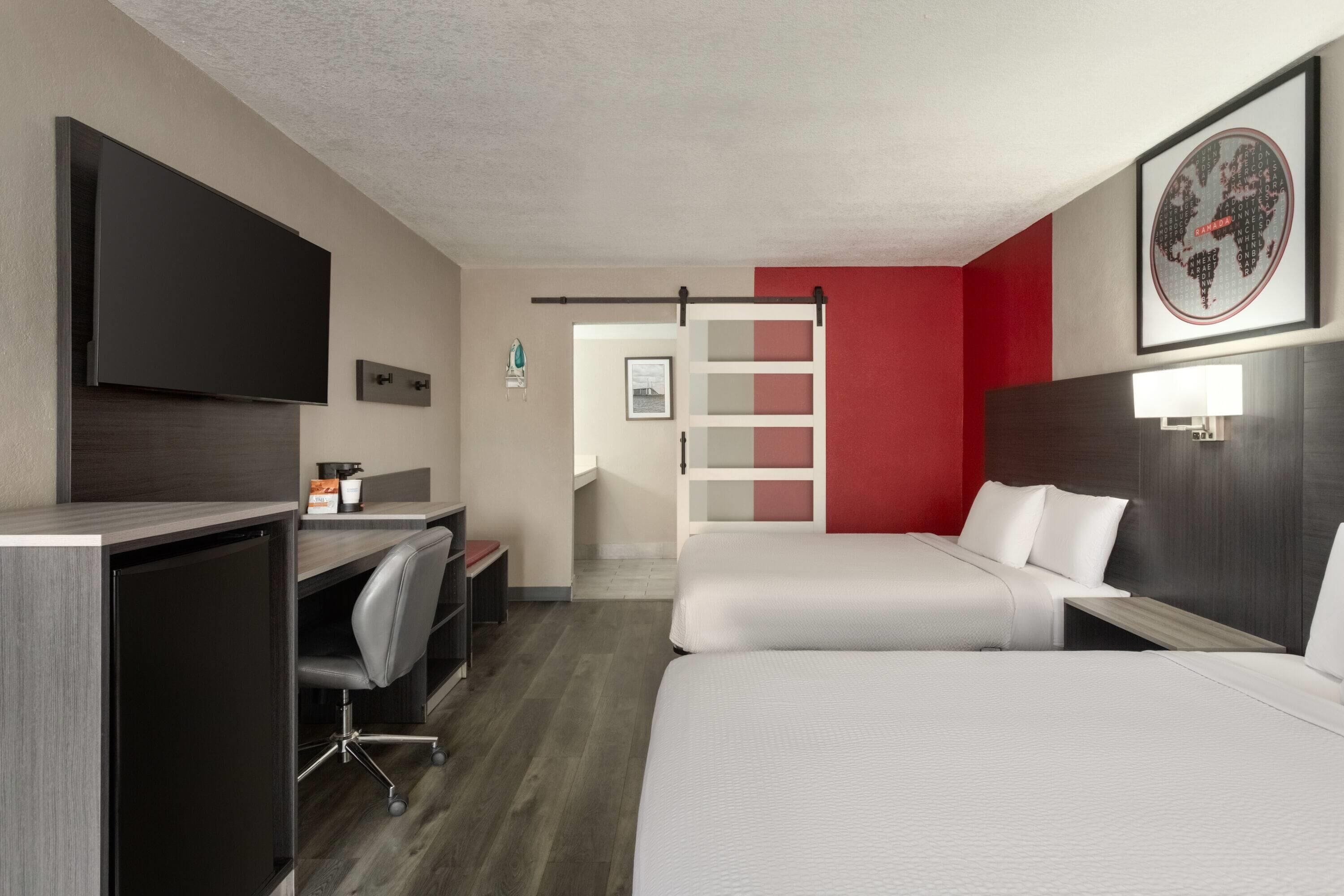 Habitación de hotel con escritorio, dos camas, televisor y una pared de acento roja.