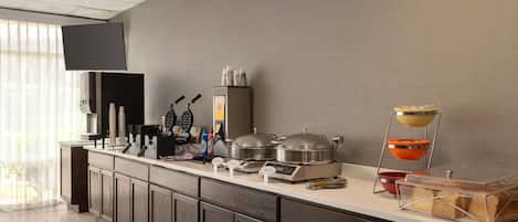 A hotel breakfast bar with a flat-screen TV, coffee machines, and a variety of food items.