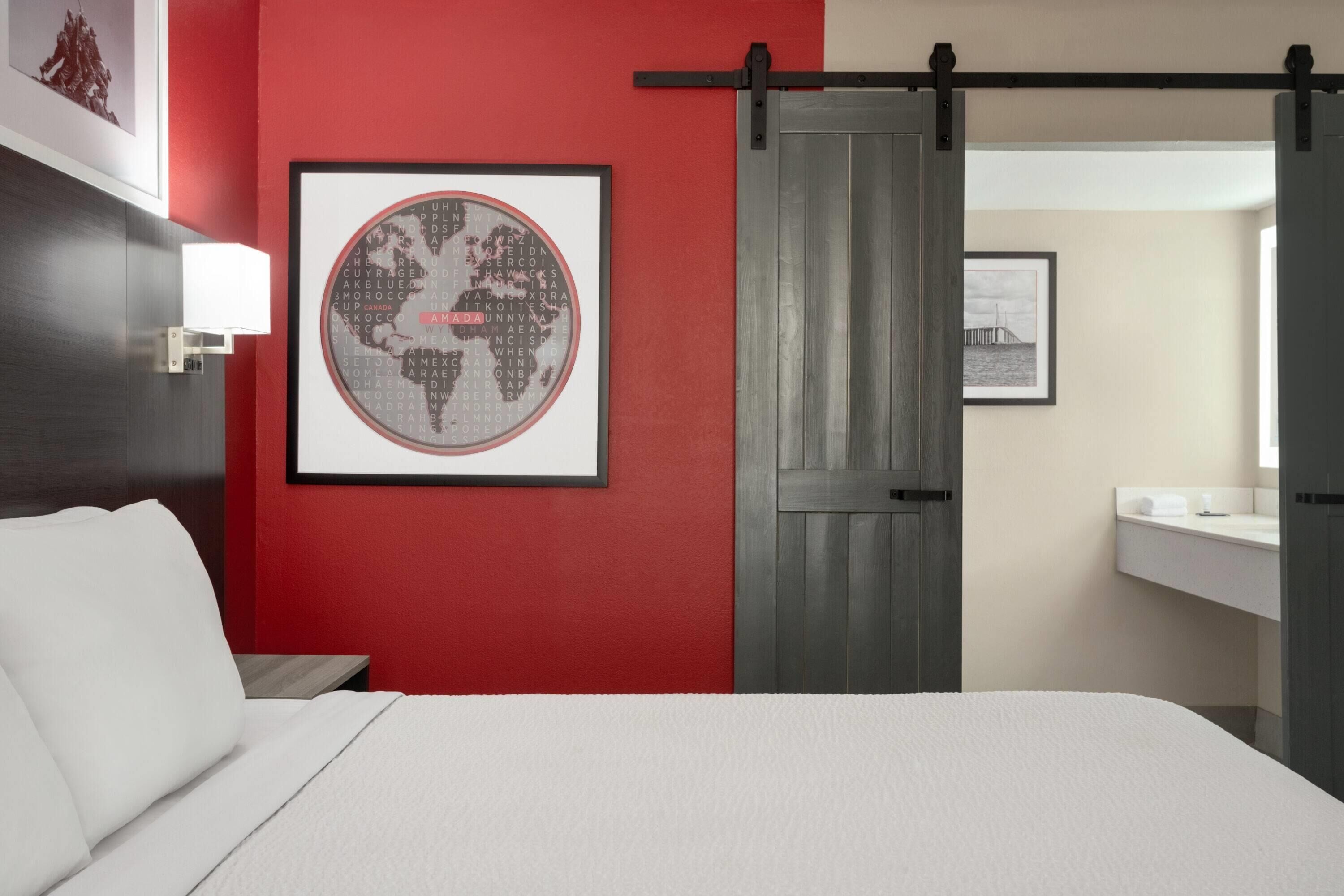 A hotel room with a bed, a red accent wall, a framed world map, a sliding door, and a bathroom with a sink and mirror.
