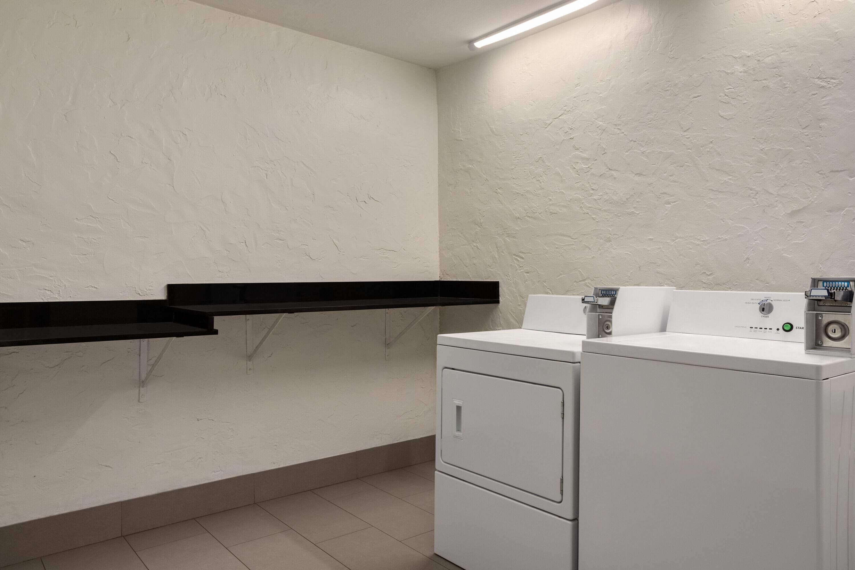 A laundry room with a washing machine and dryer, a black shelf, and a wall-mounted light fixture.