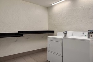 Un lavadero con lavadora y secadora, un estante negro y una lámpara fijada a la pared.