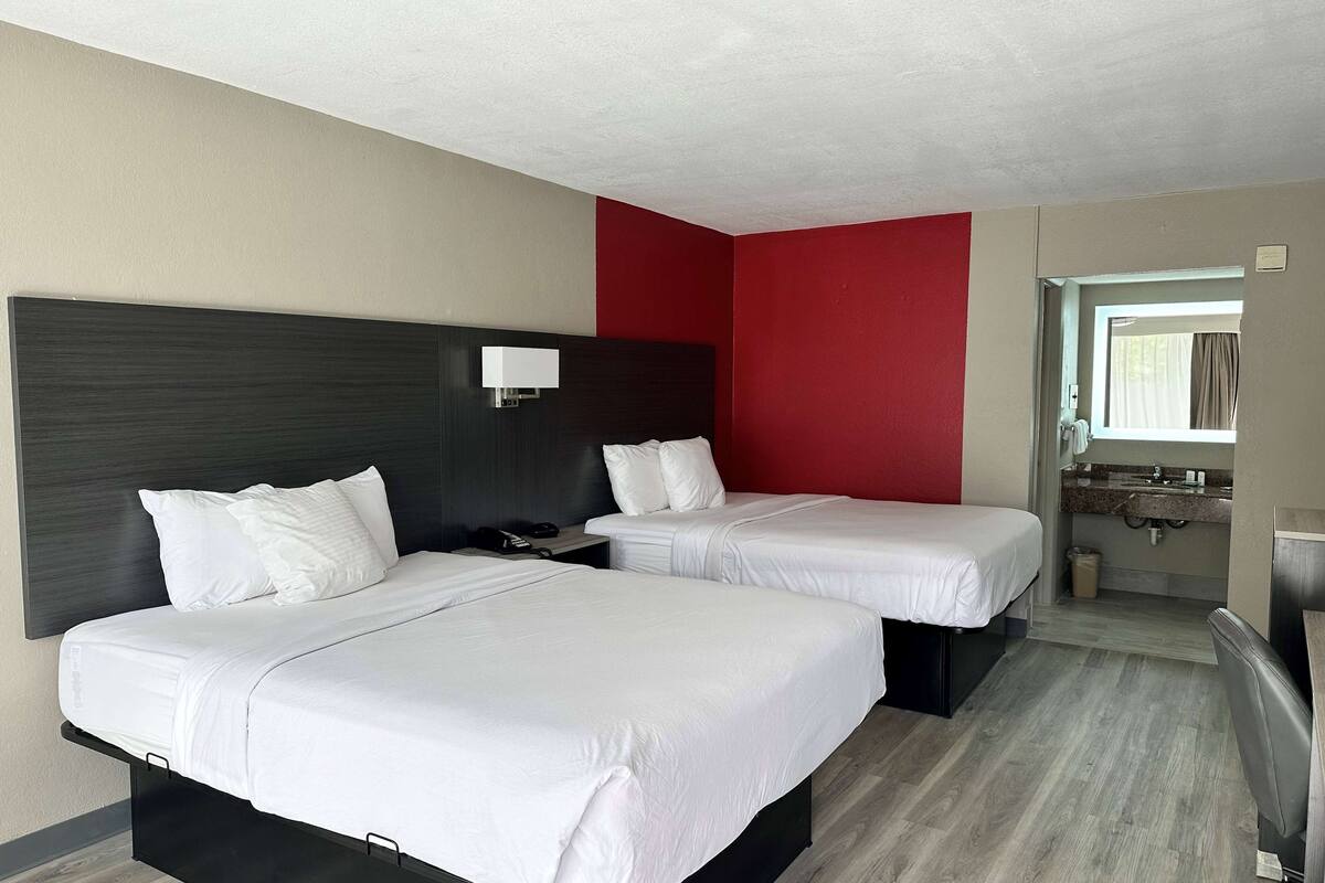 A hotel room with two beds, a red accent wall, a bathroom with a sink, and a window.