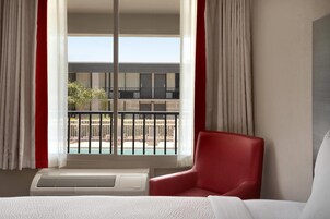 Habitación de hotel con cama, un sillón rojo, una ventana que ofrece vista a una piscina y un balcón, y aire acondicionado en el techo.