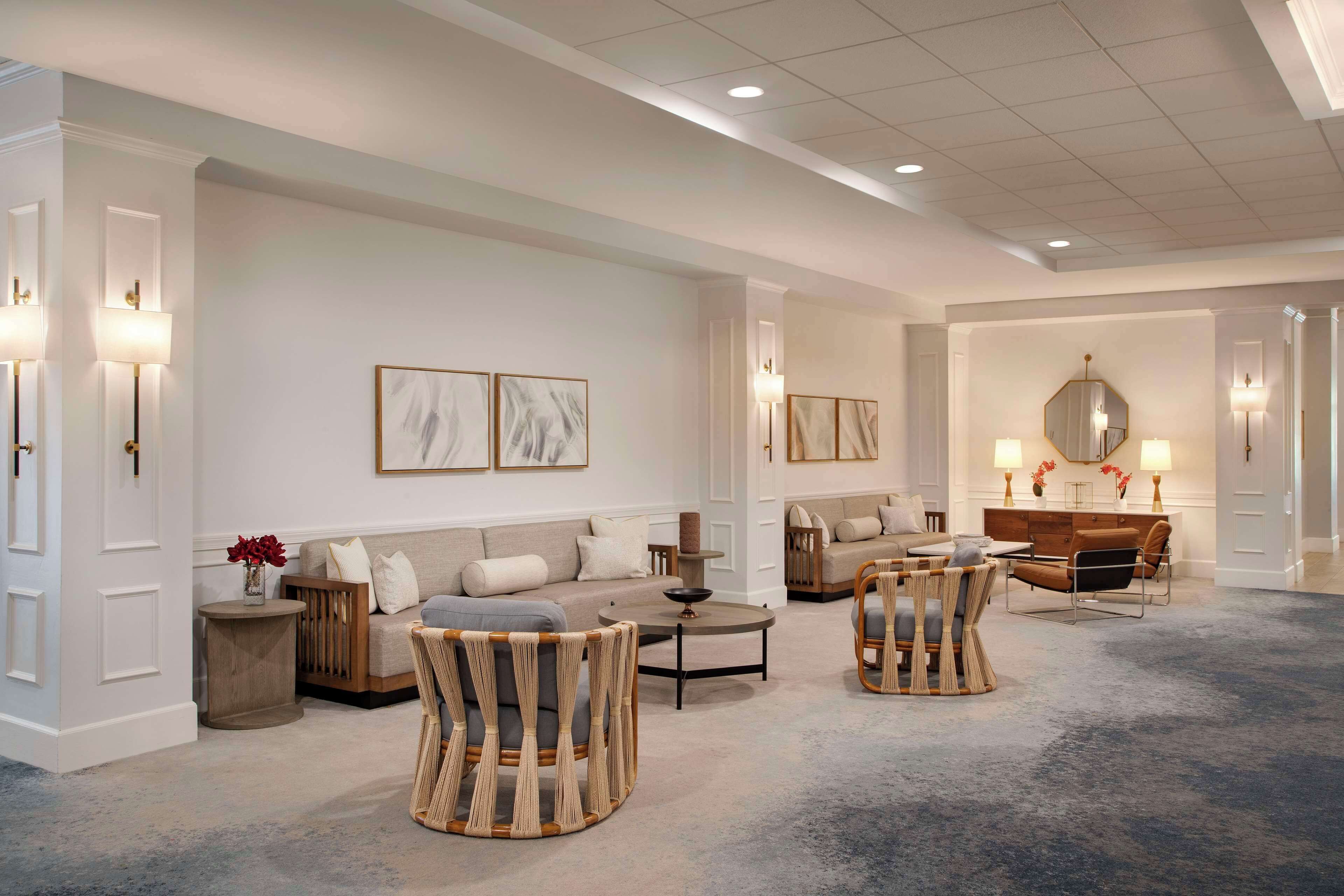 A spacious hotel lobby with a neutral color palette, featuring a seating area with sofas and chairs, a coffee table, and a decorative mirror.